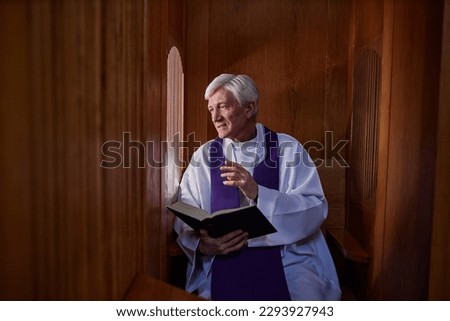 Senior priest reading prayer from Bible while talking to sinner during confession in booth Royalty-Free Stock Photo #2293927943