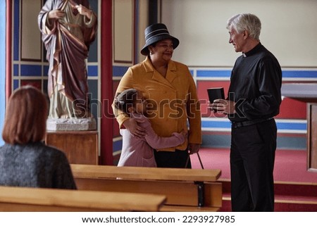 Grandmother with her granddaughter talking to priest while standing in church after ministration