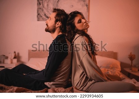 Displeased man sitting back to back with girlfriend on bed in evening Royalty-Free Stock Photo #2293934605