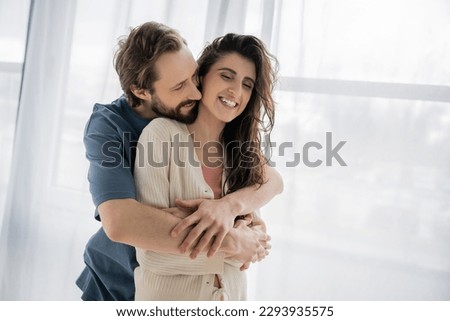 Bearded man embracing cheerful brunette girlfriend at home Royalty-Free Stock Photo #2293935575