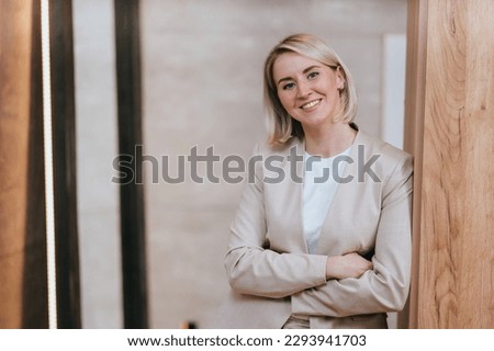 Blonde caucasian young woman in beige suit standing at office looks at camera toothy smiles leans  on wooden column. Cheerful American business woman satisfied by career. Successful people.