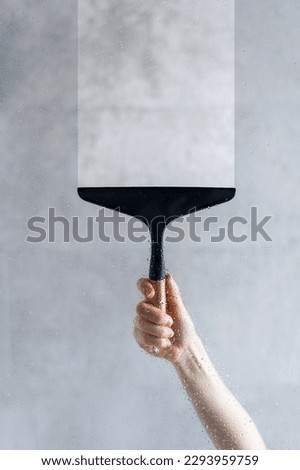 Close up view of woman hand cleaning wet shower partition by black glass wiper on the grey blurred background in the bathroom. Dry, clean, transparent and copy space stripe on the glass Royalty-Free Stock Photo #2293959759