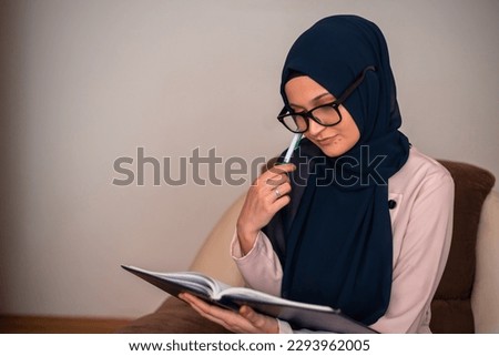 A young Muslim woman in a hijab is studying at home, writing, typing with a pencil on a notebook.