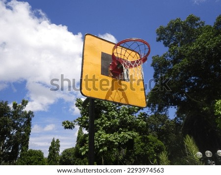 Orange basketball hoop and yellow paint board with blue sky and white clouds background on sunny day, trees around.