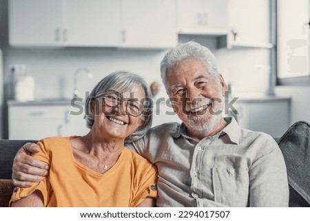 Headshot portrait of smiling elderly 60s husband and wife sit relax on couch hugging cuddling, happy mature old couple rest on sofa in living room embrace look at camera show love and care