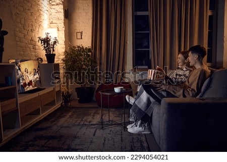 Young beautiful couple, man and woman spending time together at home in evening, sitting on couch, watching tv and eating popcorn. Concept of relationship, leisure activity, weekends, fun, emotions Royalty-Free Stock Photo #2294054021