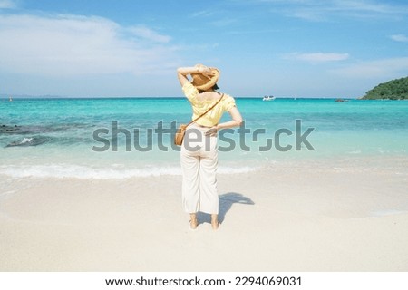 rear view of young asian woman in yellow blouse, beige pants and straw hat with sea, white cloud and blue sky as background. Summer travel. Happy tourist. Vacation on the beach. authentic people.