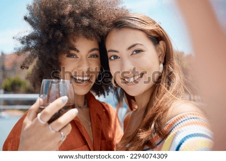 Two beautiful young mixed race women standing together and taking a selfie while bonding outside. African american with an afro drinking wine with her asian friend. Capturing memories for social media