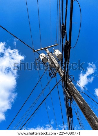 Looking upwards at a typical utility pole on a sunny day, with blue skies and white clouds. An airplane is in flight in the shot.