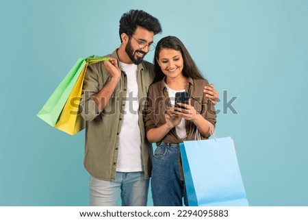 Happy cheerful beautiful young indian couple woman and man holding purchases, looking at smartphone screen and smiling, using nice shopping mobile app, blue background, copy space