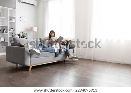 Candid shot of happy young eastern couple chilling on couch at home, using gadgets smartphone and digital tablet, indian man and woman relaxing at weekend together, copy space, full length Royalty-Free Stock Photo #2294095913