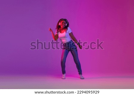 Feel The Beat. Carefree Black Woman In Wireless Headphones Dancing In Neon Light, Cheerful Young African American Woman Making Energetic Moves And Having Fun Over Purple Studio Background, Copy Space