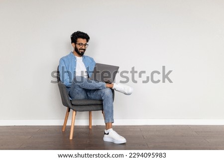 Freelance Career. Smiling Indian Man Using Laptop Computer While Sitting In Chair Over White Wall Background, Eastern Guy Browsing Internet And Working Online Online, Enjoying Remote Job, Copy Space