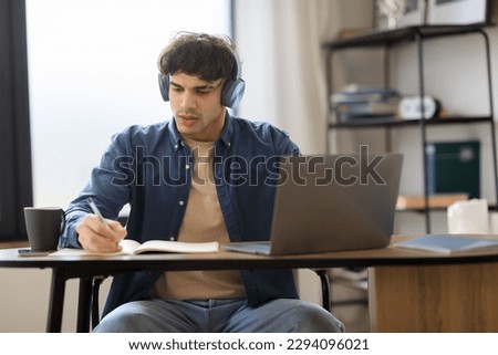 E-Learning. Middle Eastern Student Guy Taking Notes Learning Online At Laptop Computer Sitting At Table And Wearing Headphones At Home. Serious Man Writing At Workplace Indoor