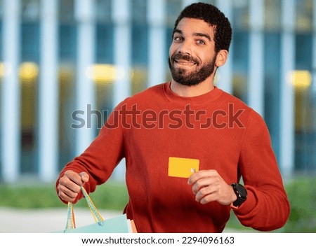 Shopping Offer. Cheerful Bearded Brunette Man Showing His Credit Card And Paper Shopper Bags, Advertising Bank Payment Service Smiling To Camera Standing Outdoors. Selective Focus
