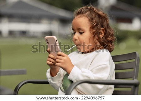 small, cute girl curly brunette, with a lollipop in her mouth and a smartphone in her hands, sitting in an outdoor chair, the concept of gadgets for children