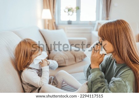 Young mother showing her small girl the right way to blow a nose. Mother helping daughter blowing her nose on the sofa at home. Adorable girl blowing nose into tissue paper by his mother at home.
