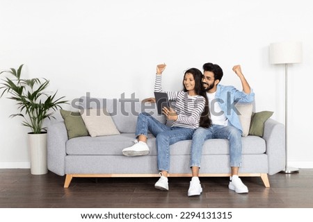 Emotional cheerful beautiful young multicultural couple middle eastern guy and indian woman sitting on sofa at home, using digital tablet and gesturing, gambling on Internet successfully, copy space