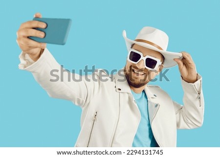 Happy man in funky fit takes selfie on phone. Cheerful attractive young guy wearing white leather jacket, panama hat and sun glasses smiles and takes photo of himself on mobile. Fashion, style concept