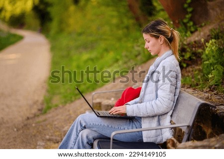 Young woman in blue coat and jeans with red backpack sitting on bench in park using laptop