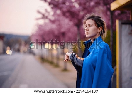 Young girl in blue jacket and jeans standing on bus stop with sakura alley looking on watch on hand Royalty-Free Stock Photo #2294164139