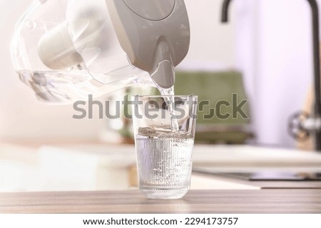Pouring of water from modern filter jug into glass on kitchen counter, closeup Royalty-Free Stock Photo #2294173757