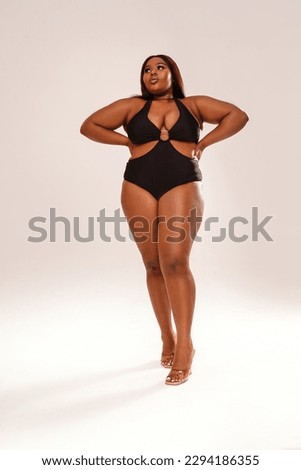 Full length photo of beautiful, dark skinned woman posing on light gray studio background, wearing black fashionable swimwear. Body positive, conscious concept. Afro hairstyle. Copy space. Selflove.
