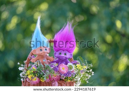 Funny toys trolls with flowers bouquet in basket close up, natural abstract green background. floral composition. spring summer season. concept of friendship, childhood. greeting card design.
