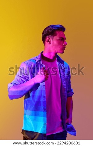 Handsome young man in casual stylish clothes posing with serious expression over yellow background with gradient neon lights. Concept of modern photography, art, cyberpunk, techno, creativity, fashion