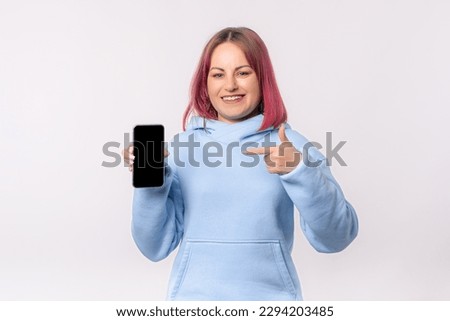 Smiling lovely woman with pink hair in a blue hoodie shows a mobile phone with a blank screen, stands over white background