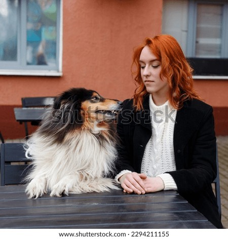 Red-haired girl with her pet sits in a cafe. Dog black and white collie breed. Similar owner and dog. Royalty-Free Stock Photo #2294211155