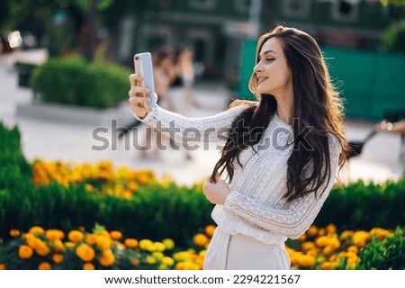 A cheerful young boho girl is standing in a city park and taking self-portraits while smiling at the phone on a sunny summer day. A fashionable woman is taking pictures of herself on a city street.
