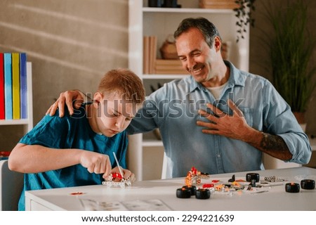 Wide shot of a blond boy assembling his metal toy with patience and concentration while his father is watching with proud and joy. Beautiful family moments between a loving father and his teenage son. Royalty-Free Stock Photo #2294221639