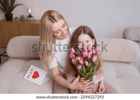 Happy Mother's Day! The child's daughter congratulates her mother and gives her flowers tulips and a card. Mom and girl smile and hug. Family vacation and togetherness.