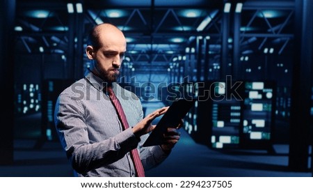 Information security officer working in data center with tablet, analyzing server rack cabinets in render farm. IT support specialist inspecting cloud computing system, ai concept. Tripod shot. Royalty-Free Stock Photo #2294237505