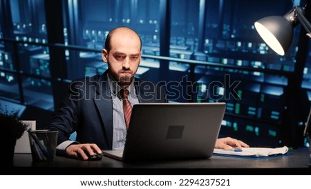Male infrastructure engineer working in render farm running diagnostics on mainframe computer system in modern data center. Database admin controlling work of server cabinets on laptop. Royalty-Free Stock Photo #2294237521