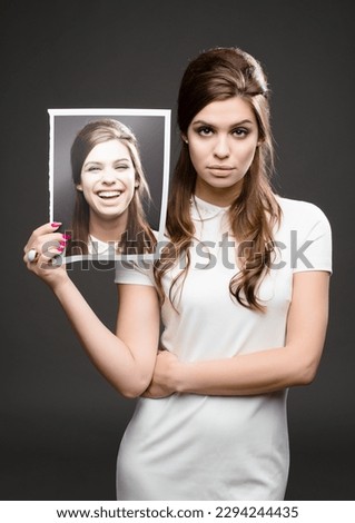 Different day, different mood. Studio shot of an attractive young woman dressed up in 60s wear and holding a photograph of herself smiling against a dark background.