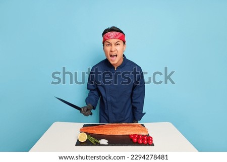 Serious cook going to slice fish fillet, holding knife in his hand, standing near the cooking table in the studio, wearing blue chef's jacket, professional people concept, copy space, high quality