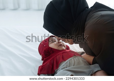 Muslim mom plays and teases happy baby daughter on bed, mother and little baby girl wears hijab, kiss the baby's nose tenderly, view from above, happy loving family concept