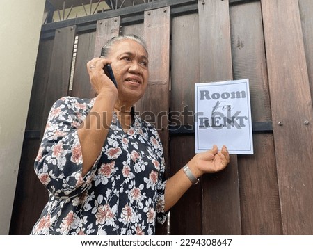 Asian Indonesian Elder Woman wearing floral dress talk holding phone front of wood gate with signboard "room for rent". Renting Room concept. Rooms for rent concept
