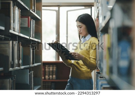 Young female college highschool student in casual outfit standing, reading or choosing books to read at library for studying, academic research, school work, project, or exam.