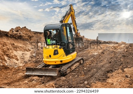 Mini excavator at the construction site on the edge of a pit against a cloudy blue sky. Compact construction equipment for earthworks. An indispensable assistant for earthworks Royalty-Free Stock Photo #2294326459