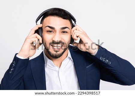 Turkish indian arab ahndsome guy man in buisness suit standing over grey background in studio isolated looking at camera holding headphones wearing receiving call call centre happy stylish man.