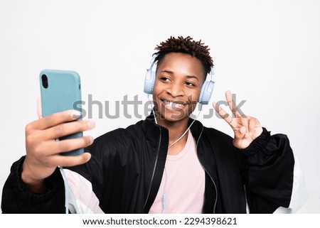 american young hipster wearing new modern wireless headphones holding cell phone smartphone in hands listening to music enjoying songs taking selfie picture shwoing two fingers at camera.
