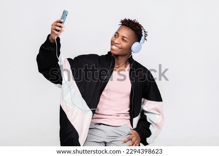 Happy dark skinned man in good mood wearing new modern wireless headphones holding cell phone smartphone in hands taking picture selfie over white background in studio isolated smiling.