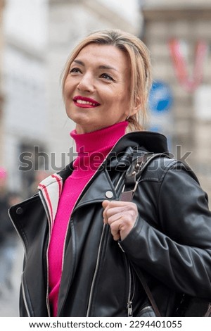 Street portrait of a smiling woman 40-45 years old in a jacket and with a bag on her shoulder on a blurry urban background.