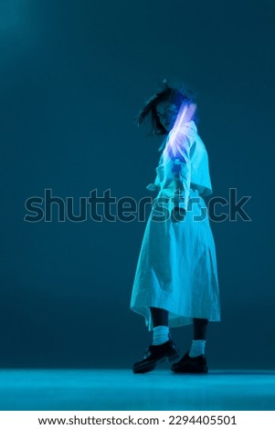 Incognito. Portrait of girl with flying hair in trendy clothes posing with neon reflection of neon glow on body over turquoise mode background. Concept of digital art, fashion, cyberpunk, futurism