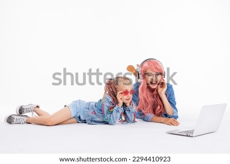 Young mom and preschooler daughter in denim clothes laying on wite background in studio waching cartoons on laptop. Mother wearing pink wig and headphones and kid with cloroful braids and sunglasses.