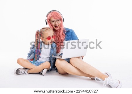 Young mother with pink wig and new modern white headphones sitting with adorable preschooler daughter on floor in white studio watching cartoons on laptop wearing denim clothes.