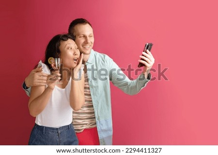 Two young people check out amazing article on web page via cellular, have surprised happy expressions, dressed in summer clothes, use wireless internet against pink background.
